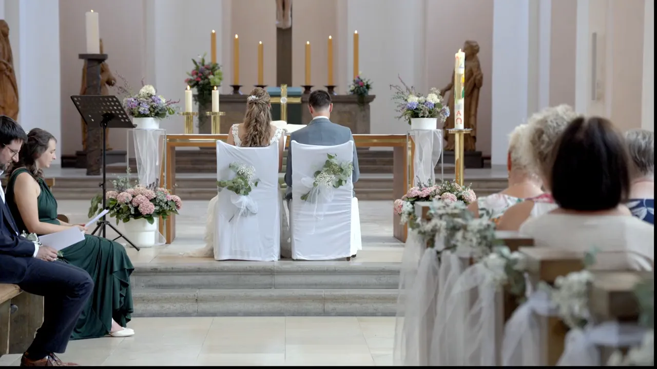 Kirchliche Trauung: das Brautpaar vor dem geschmückten Altar, Ringtausch und Kuss vor den Gästen in den Bänken.